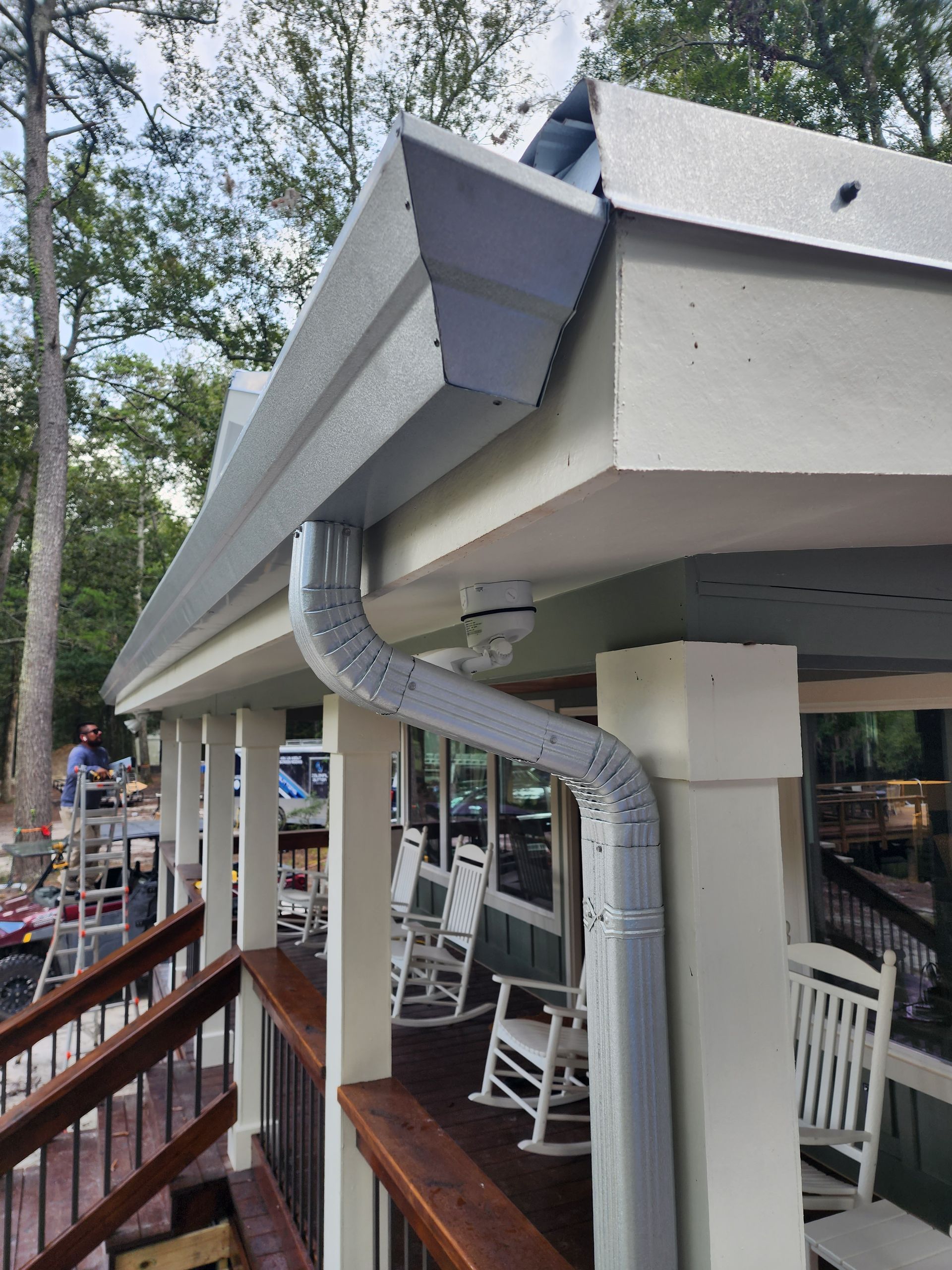 A building's corner with gray gutters, a downspout, and a deck with white rocking chairs and a wooden railing.