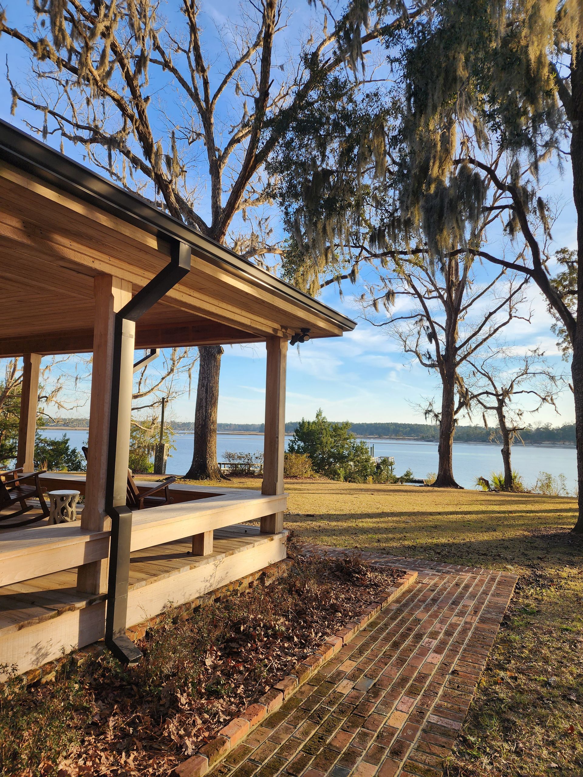 Wooden pavilion overlooking a lake, with trees and a brick path on a sunny day.
