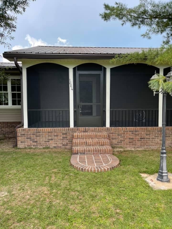 Screened porch with brick steps and black screen windows. Brown metal roof. Green grass and light post in front.