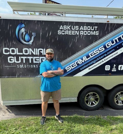 Man in blue shirt and khaki shorts stands in front of a trailer advertising seamless gutters.