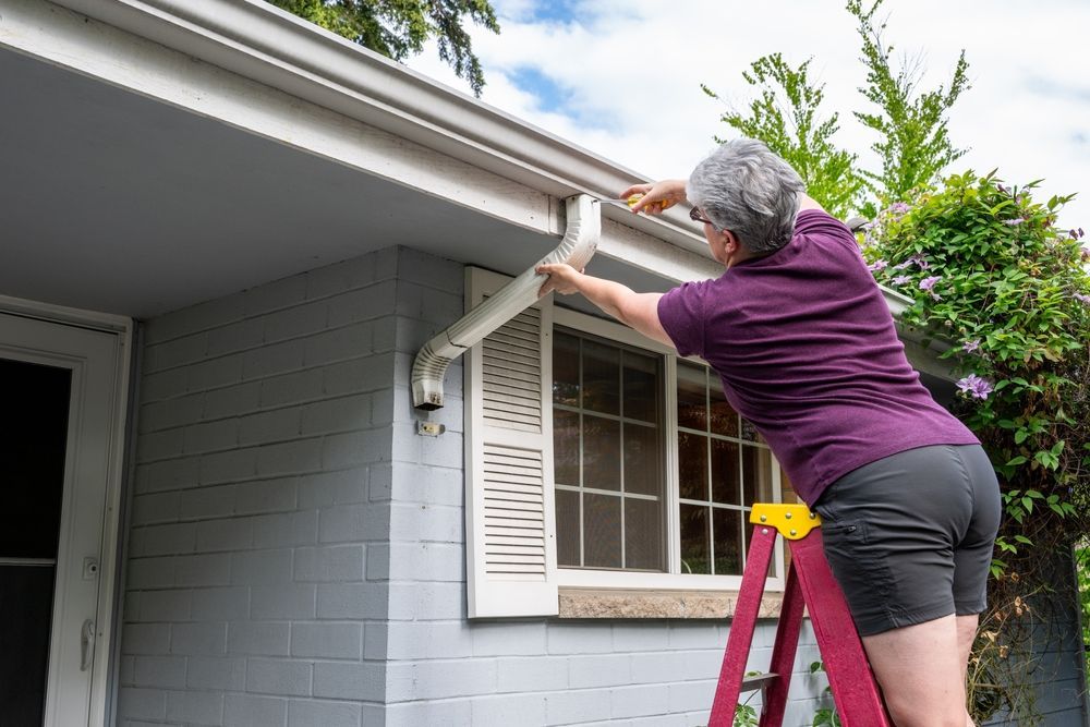 A woman is standing on a ladder fixing a gutter on a house.