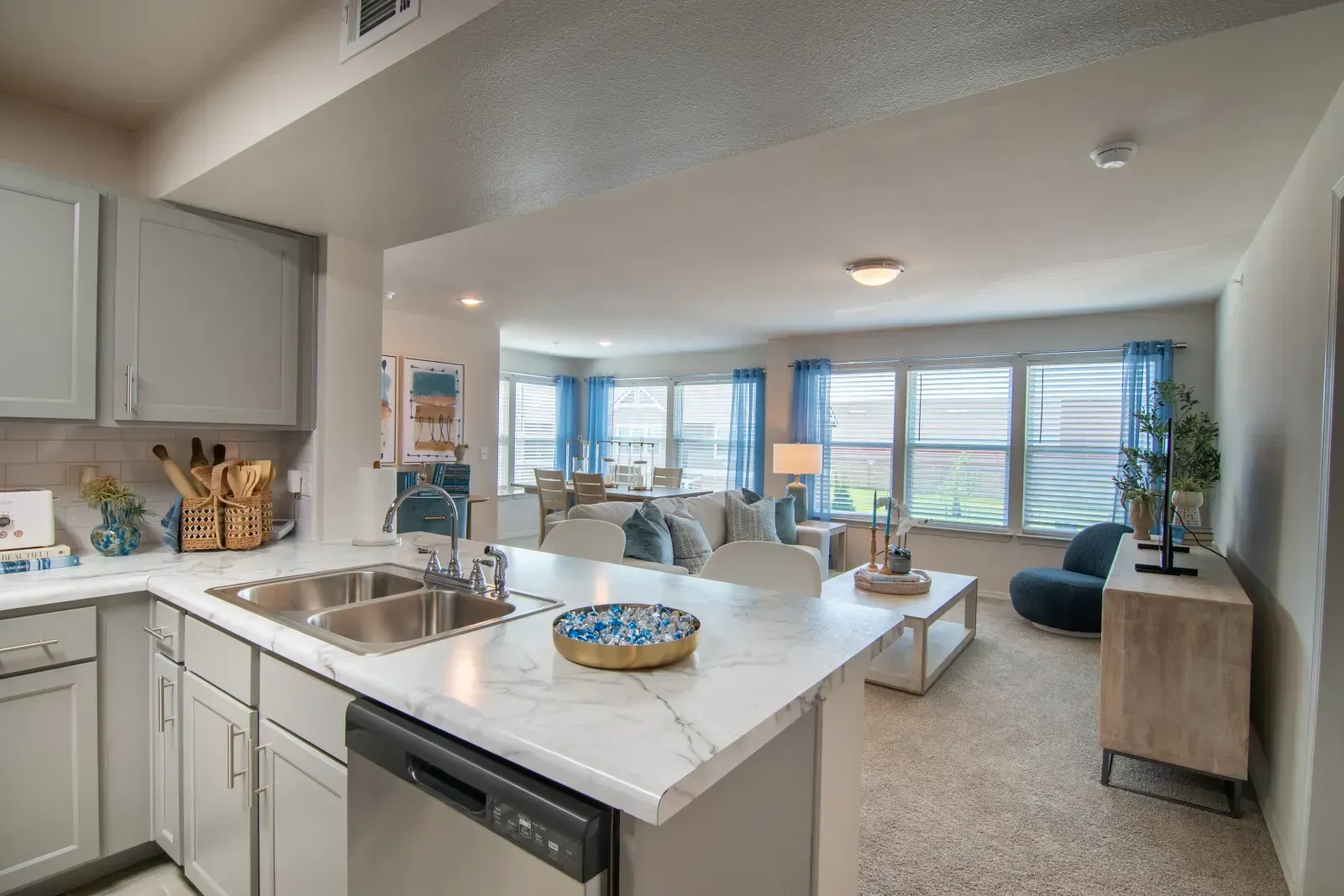 Open-concept kitchen and living area with marble countertops and blue-curtained windows.