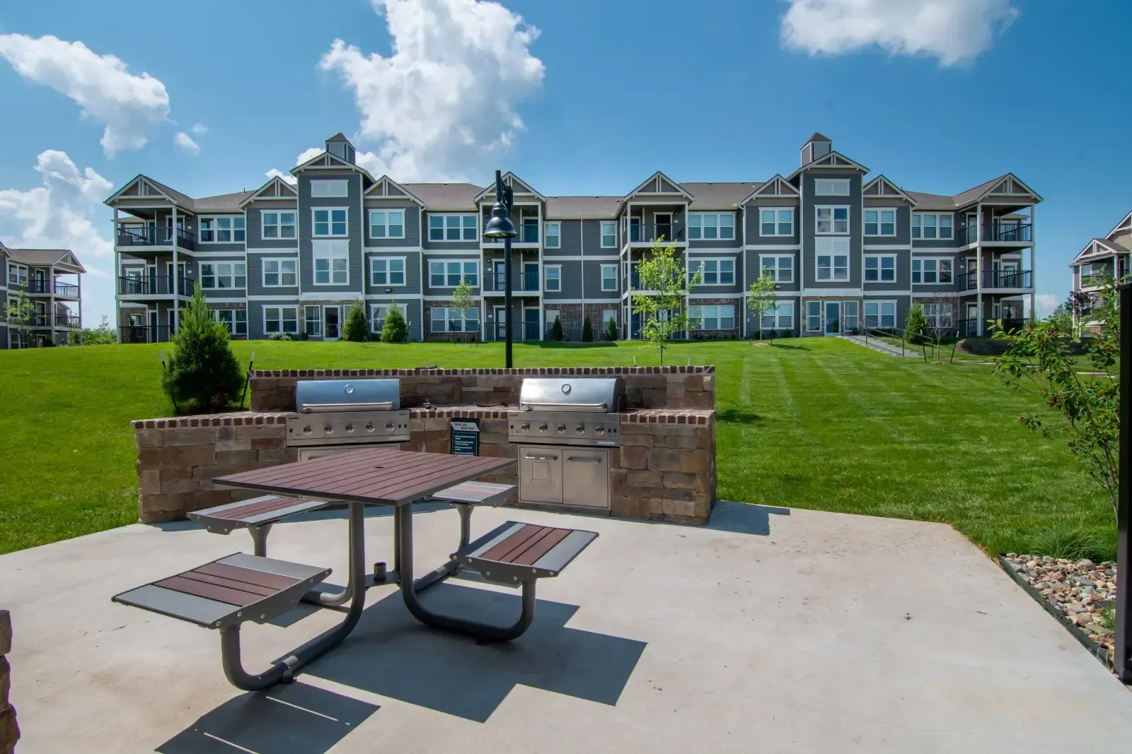 Outdoor communal grilling station with a picnic table in front of a multi-building apartment complex.