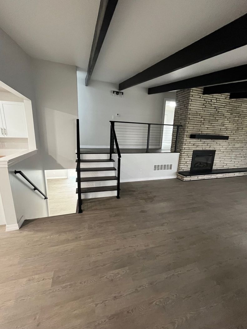 Modern living room with wood floors, stone fireplace, black beam ceiling, and a stairway nook