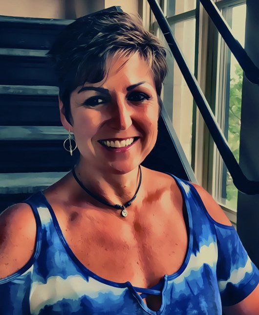 A woman in a blue tie dye shirt is smiling in front of stairs