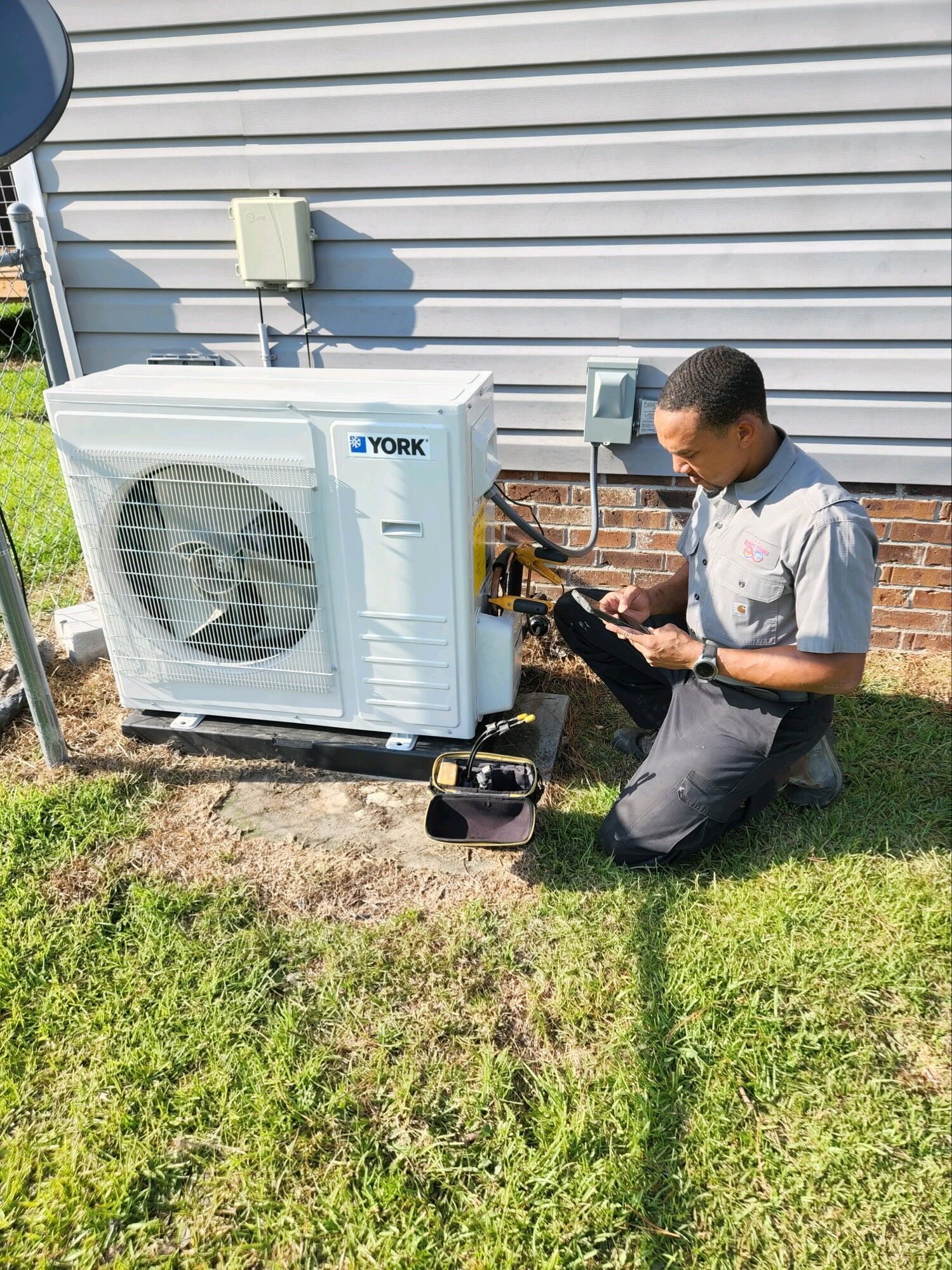 Technician kneeling beside a wall-mounted outdoor AC unit, checking wiring on a sunny lawn.