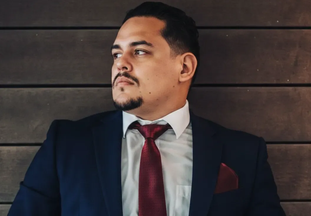 A person in a dark suit, white shirt, and maroon tie and pocket square looks away against a dark horizontal wood wall.