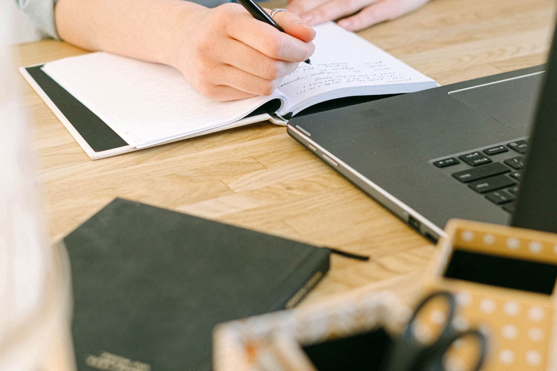 An image of someone writing in a notebook with a computer in front of them.