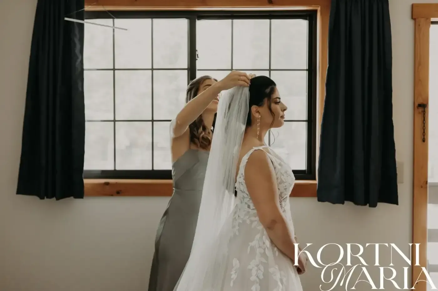 A woman is helping a bride with her veil in front of a window.