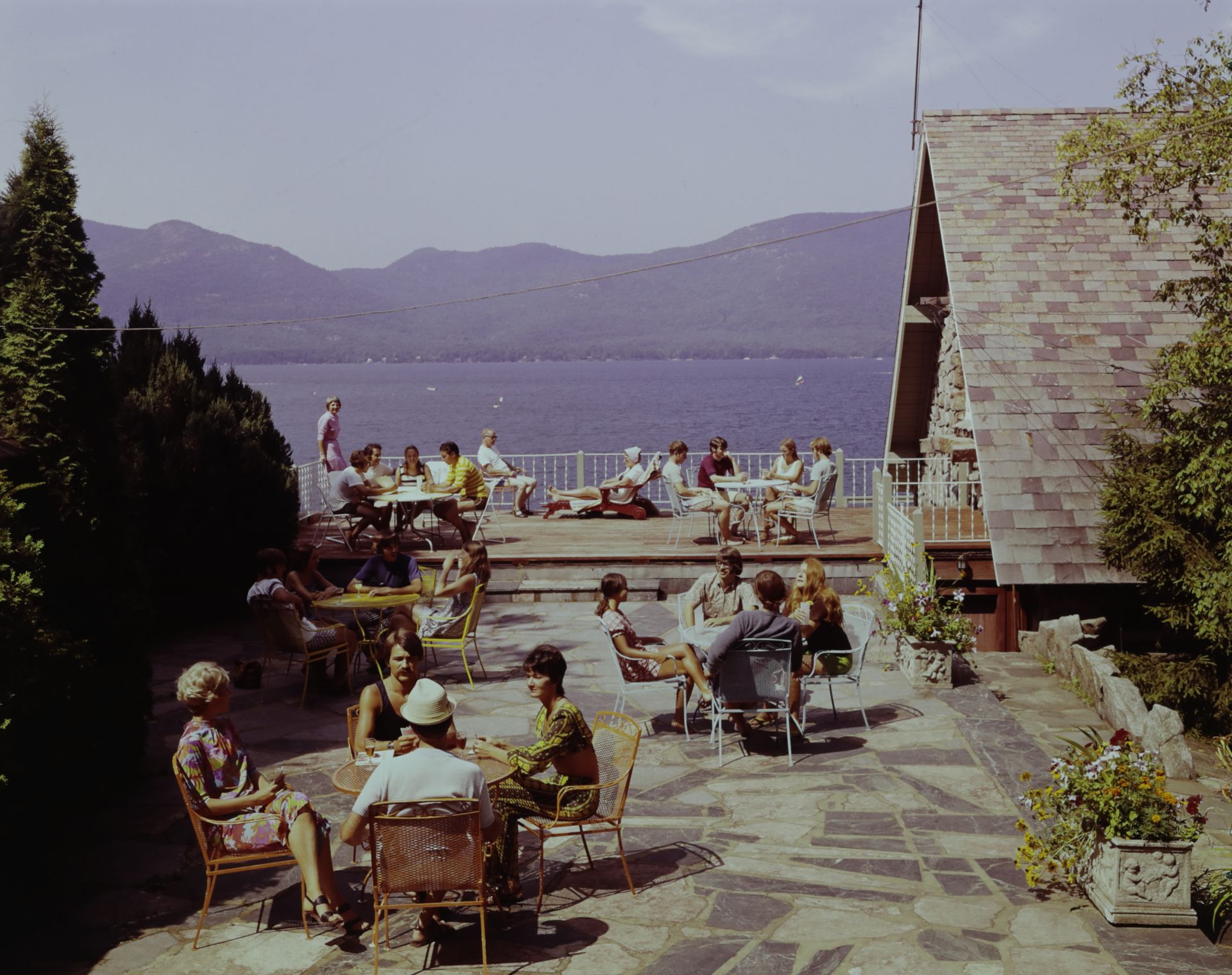 A group of people sit at tables on a patio overlooking a lake