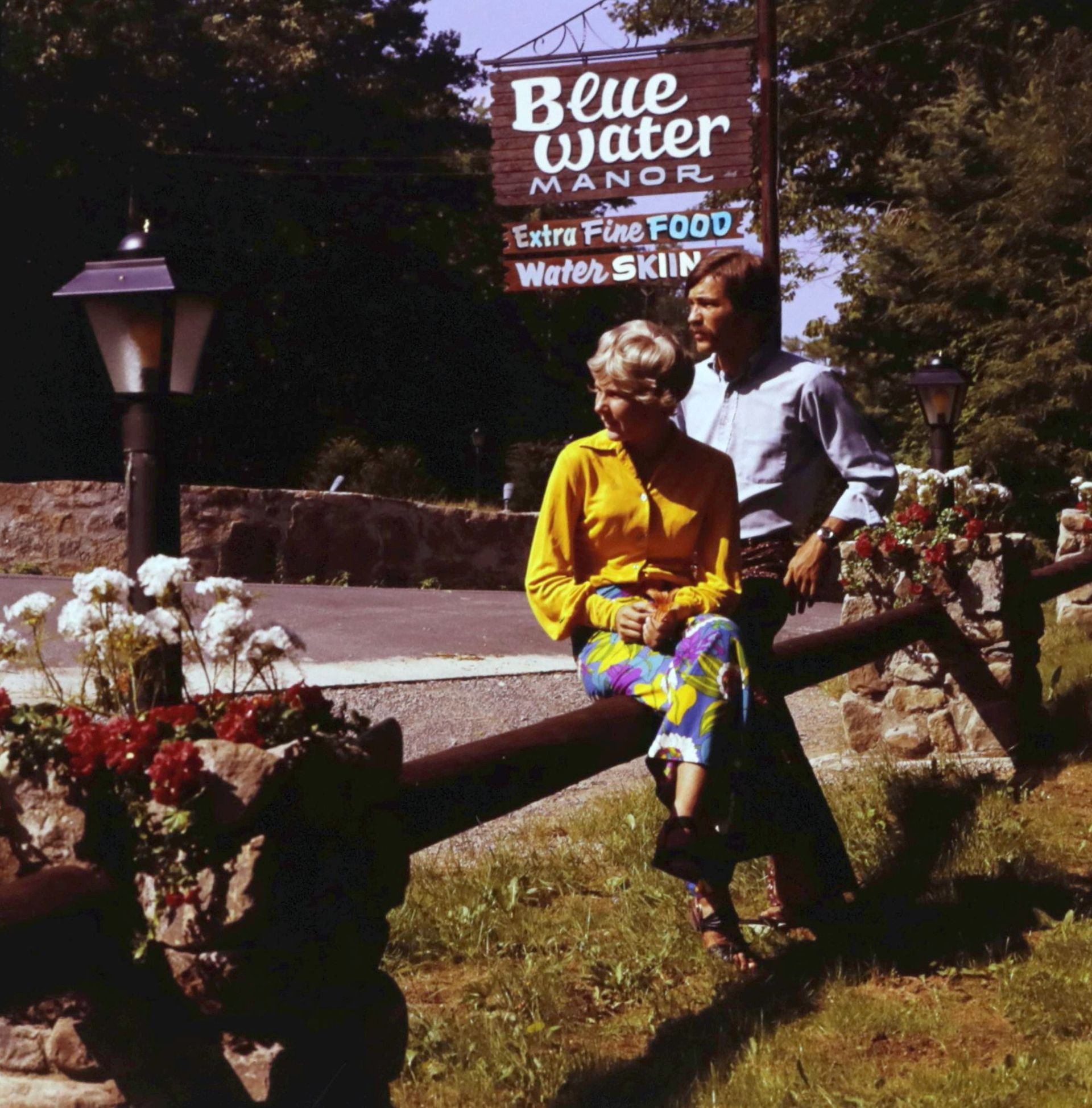 A man and woman sit on a fence in front of a blue water manor sign