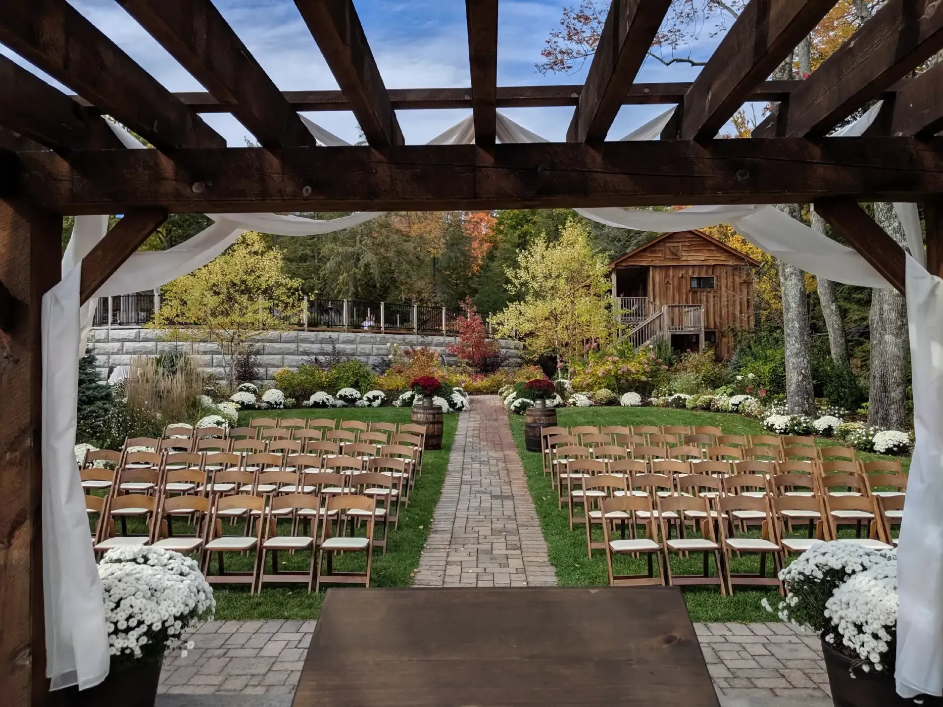 A row of wooden chairs are lined up for a wedding ceremony under a pergola.