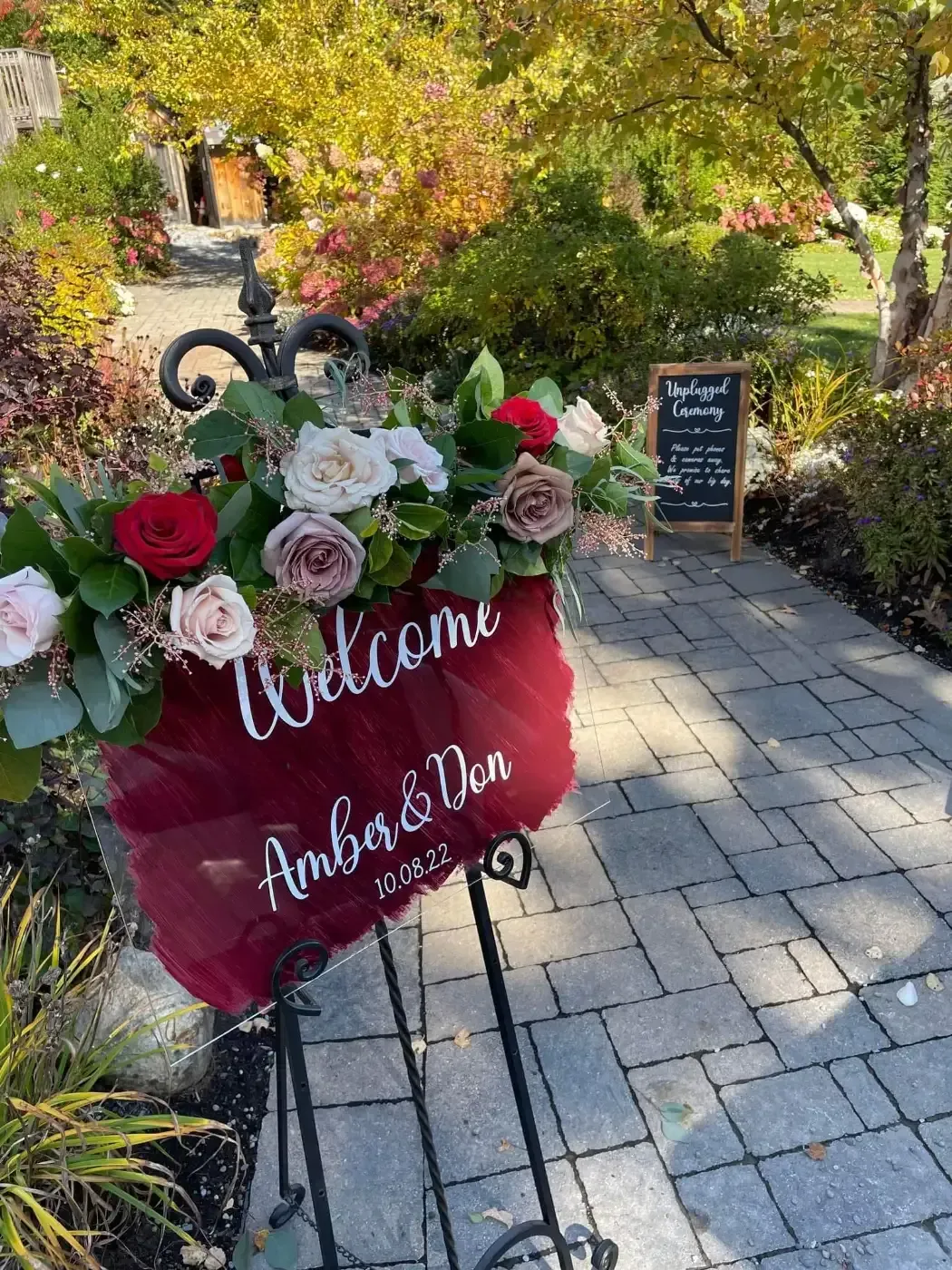 A welcome sign with flowers on it is sitting on a brick walkway.