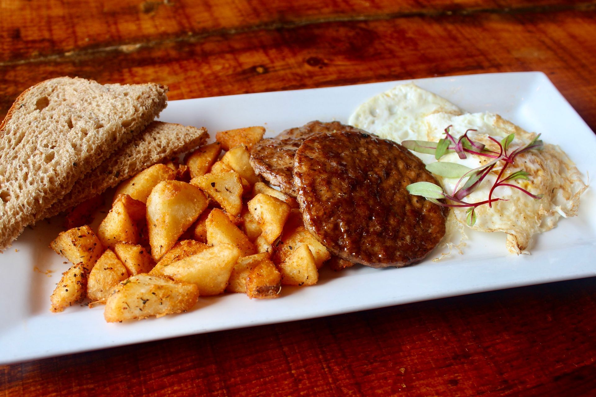 A white plate topped with meat , potatoes , eggs and toast on a wooden table.