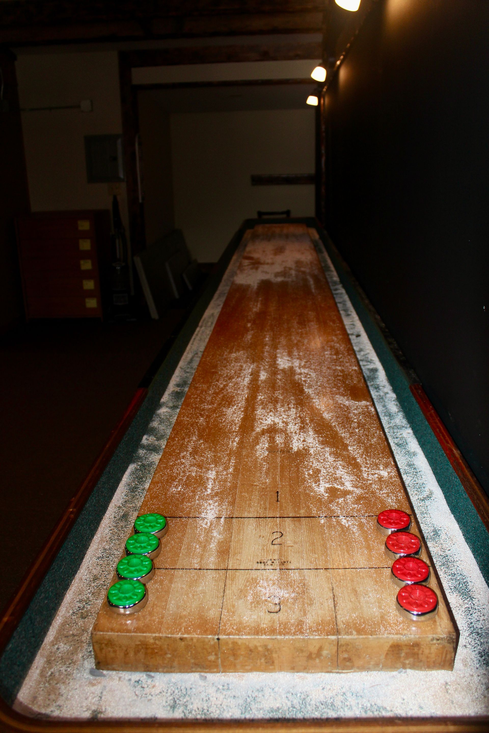 A long wooden shuffleboard table with green and red tokens on it.
