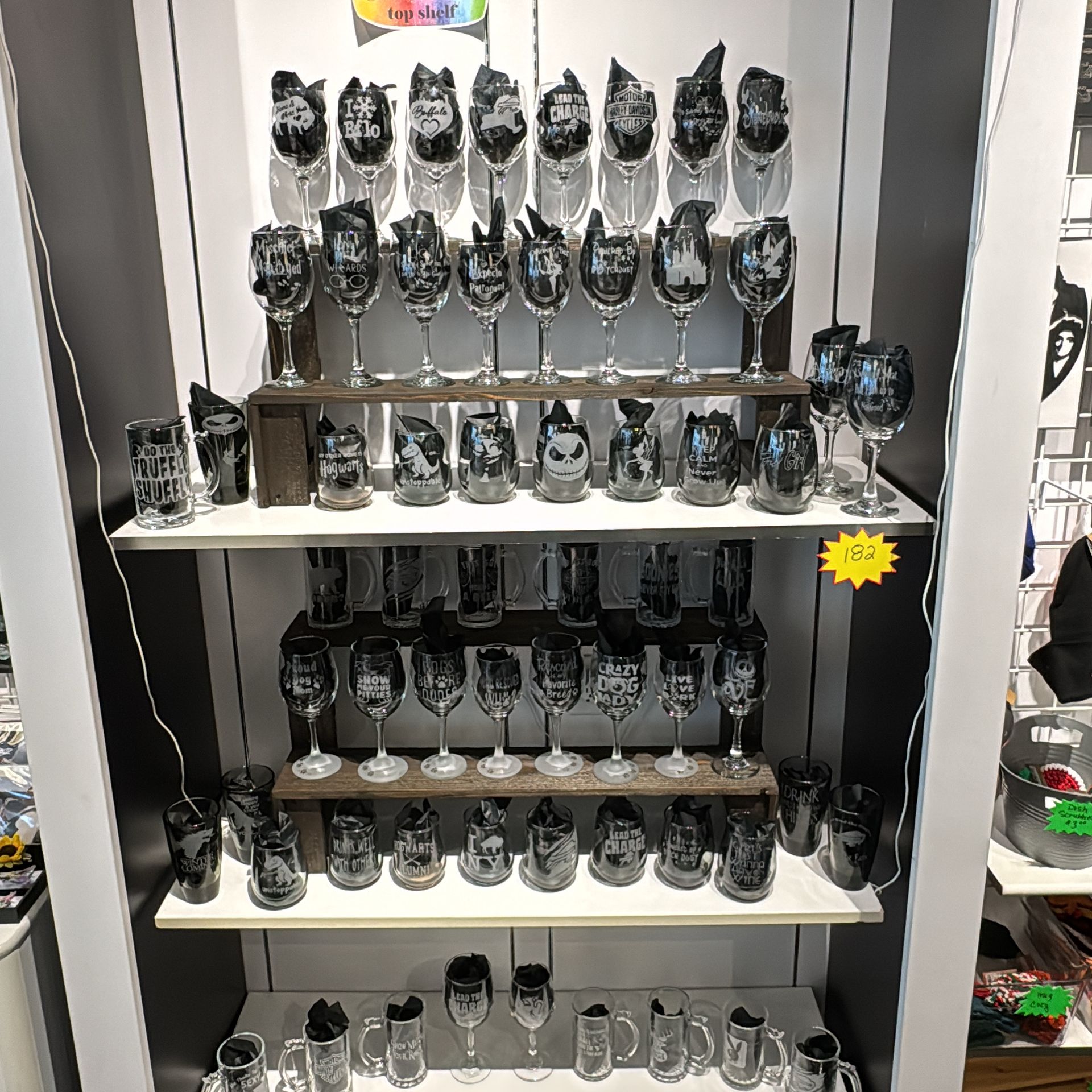 Display of engraved glassware, including wine glasses and mugs, on tiered shelves in a store.