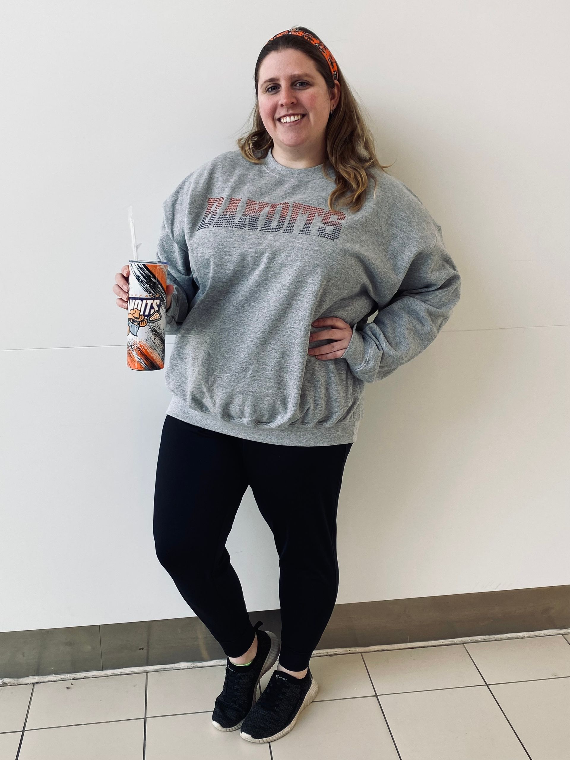 Woman in gray sweatshirt and black leggings holds a drink, smiling, by a white wall.
