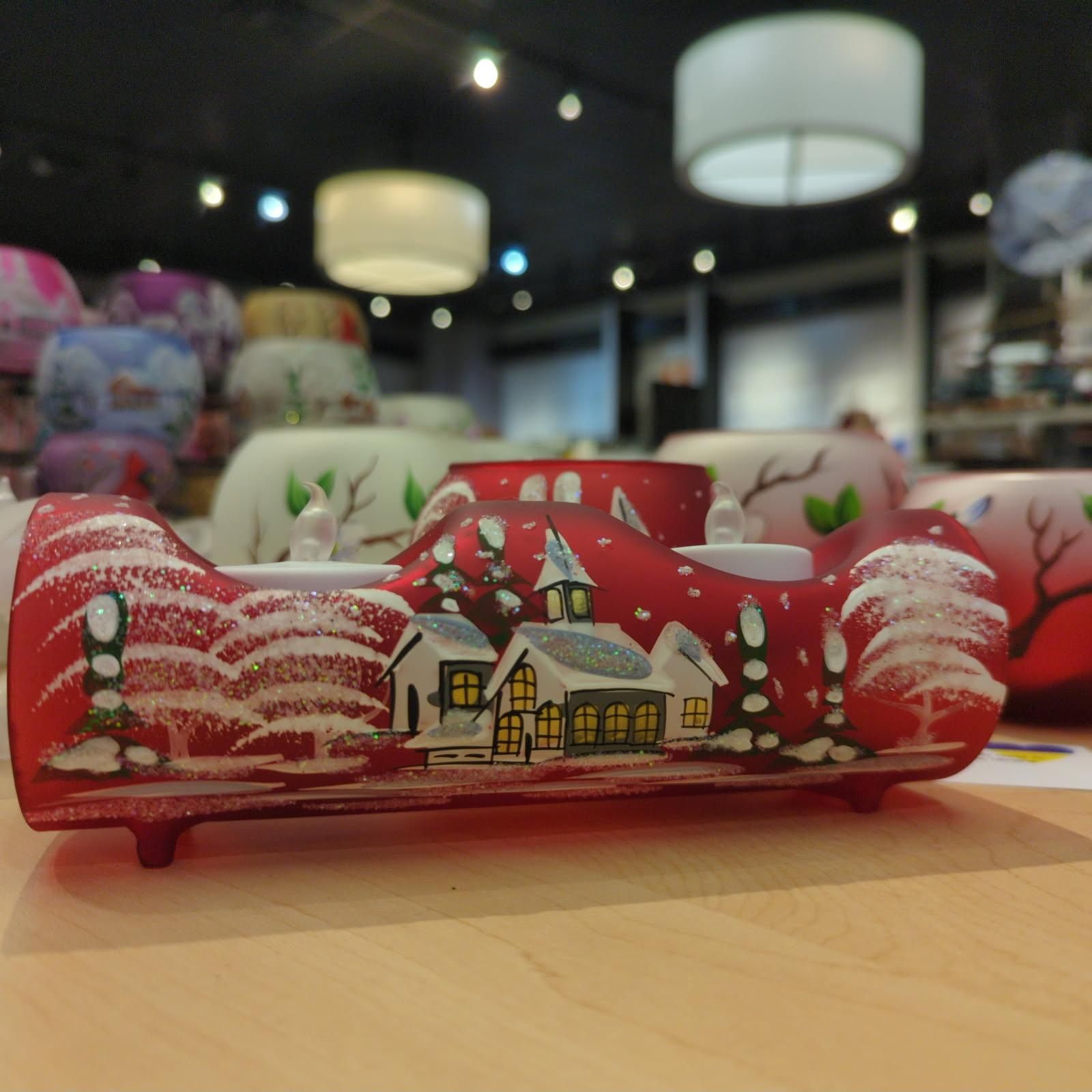 Red frosted glass candle holder, winter village scene, snow-covered trees, church, shop setting, lights in the background.