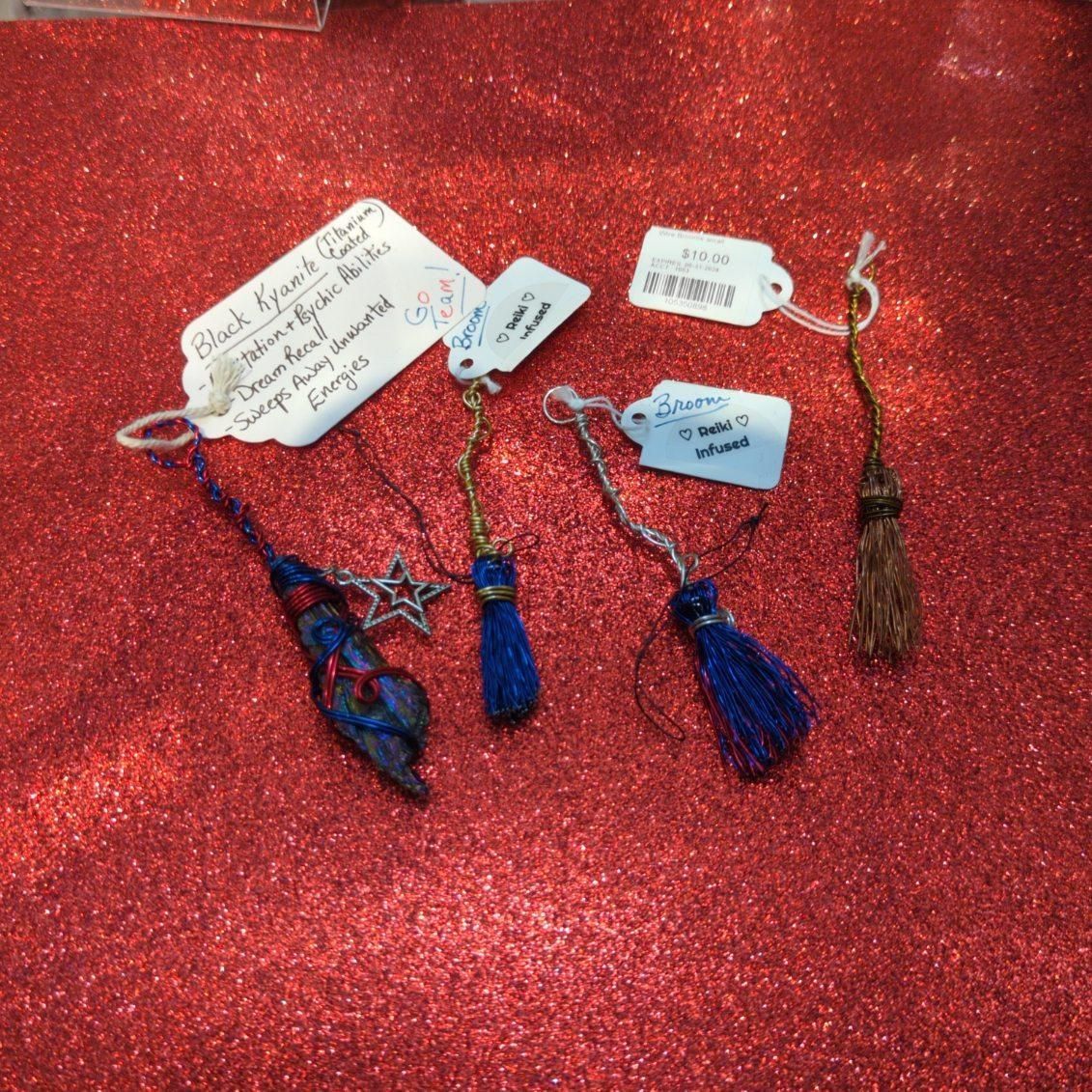 Five miniature broom decorations on a red glitter surface; each has a price tag.