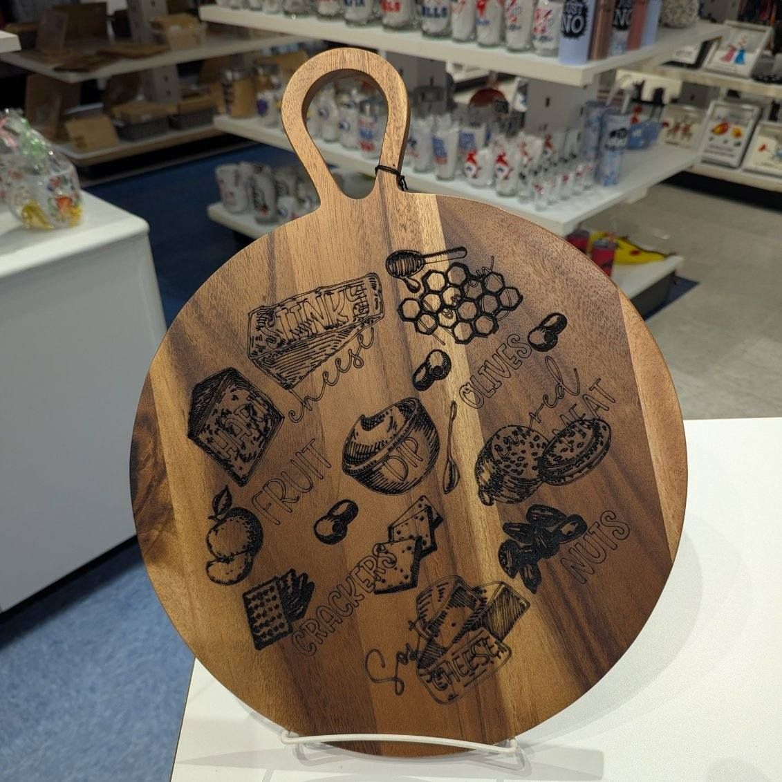 Round wooden cutting board with etched food designs. Handle at top, displayed on stand.