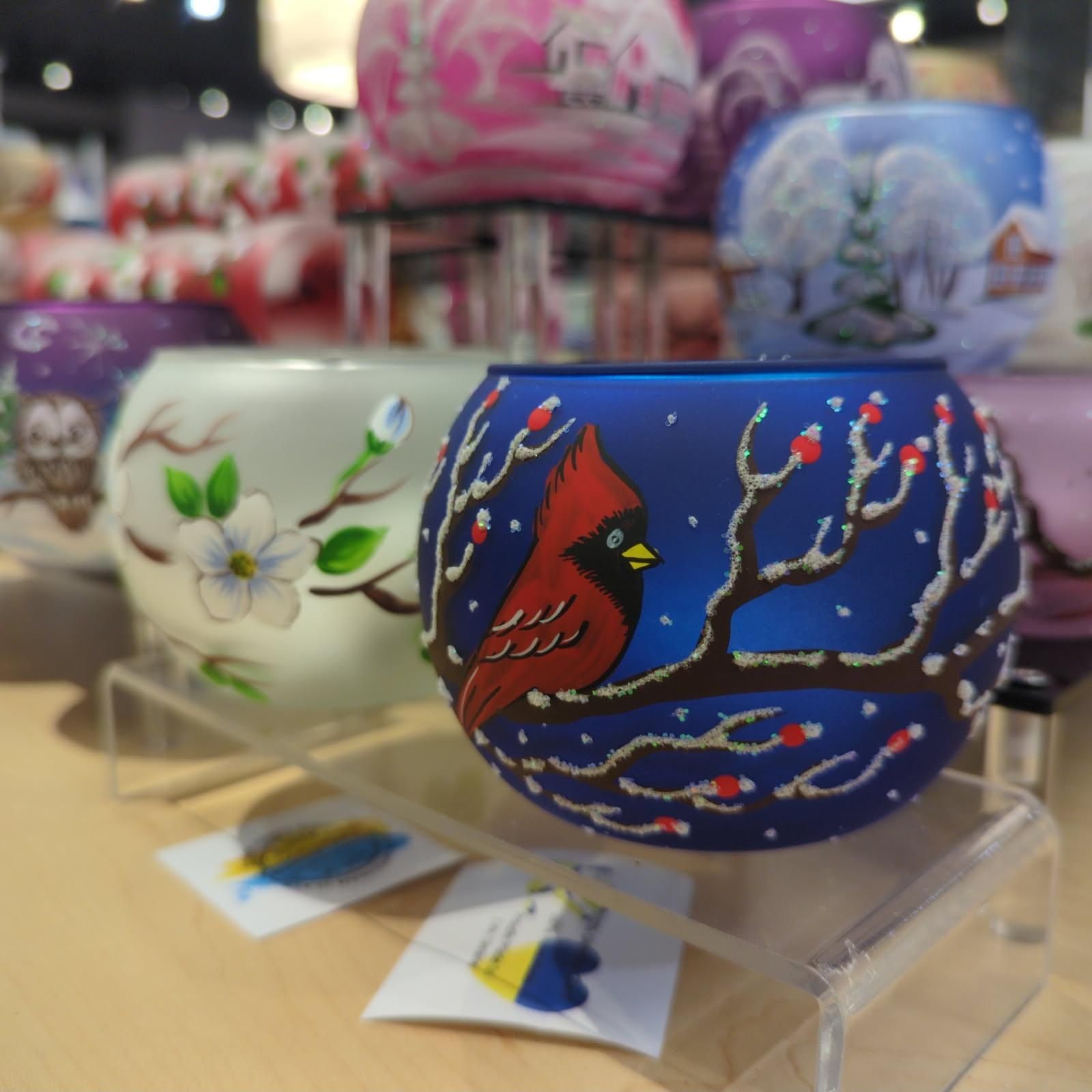 A display of round, painted glass candle holders. A blue one features a cardinal bird in a winter scene.