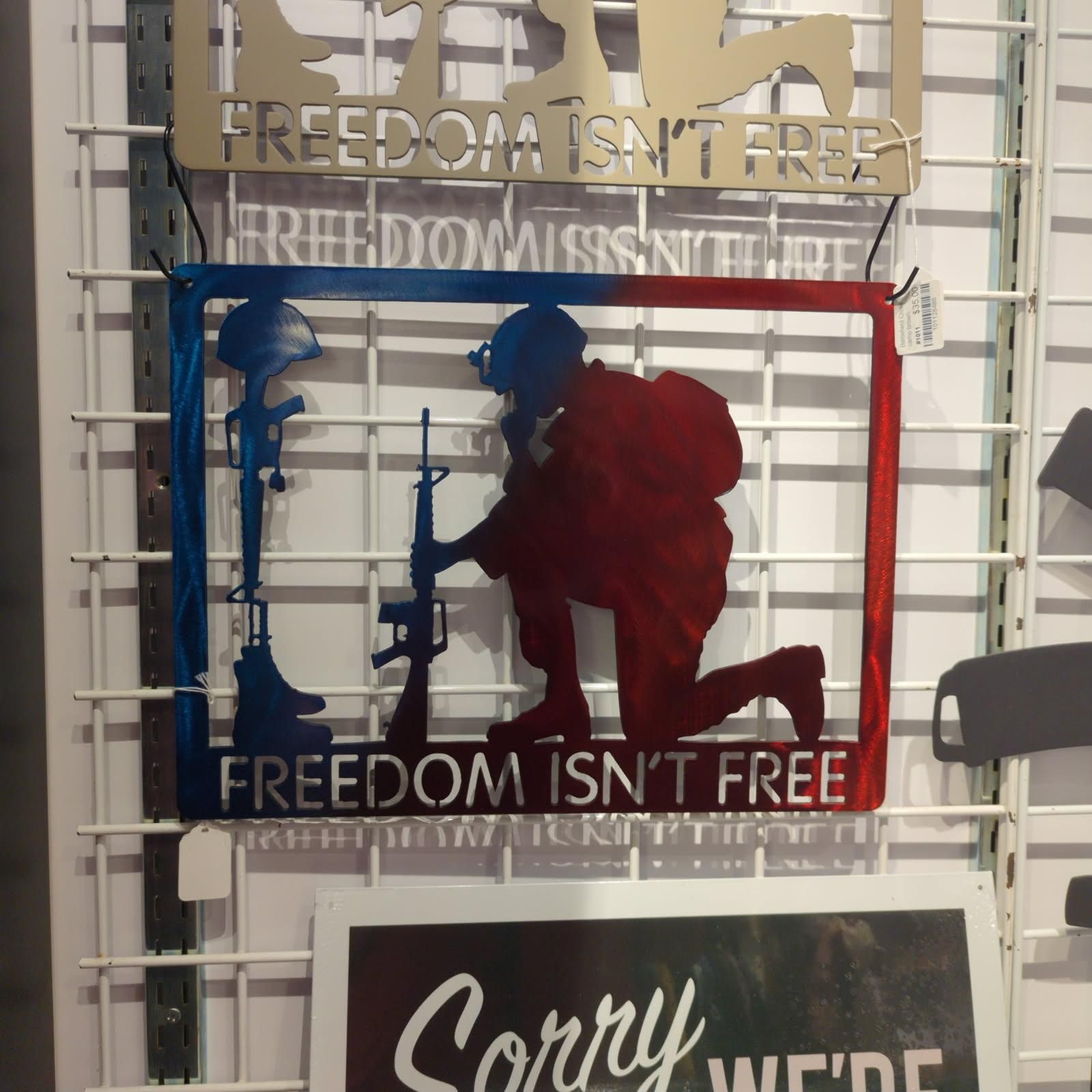 Metal sign of soldier kneeling, tombstone, rifle, in red, white, & blue, with text 