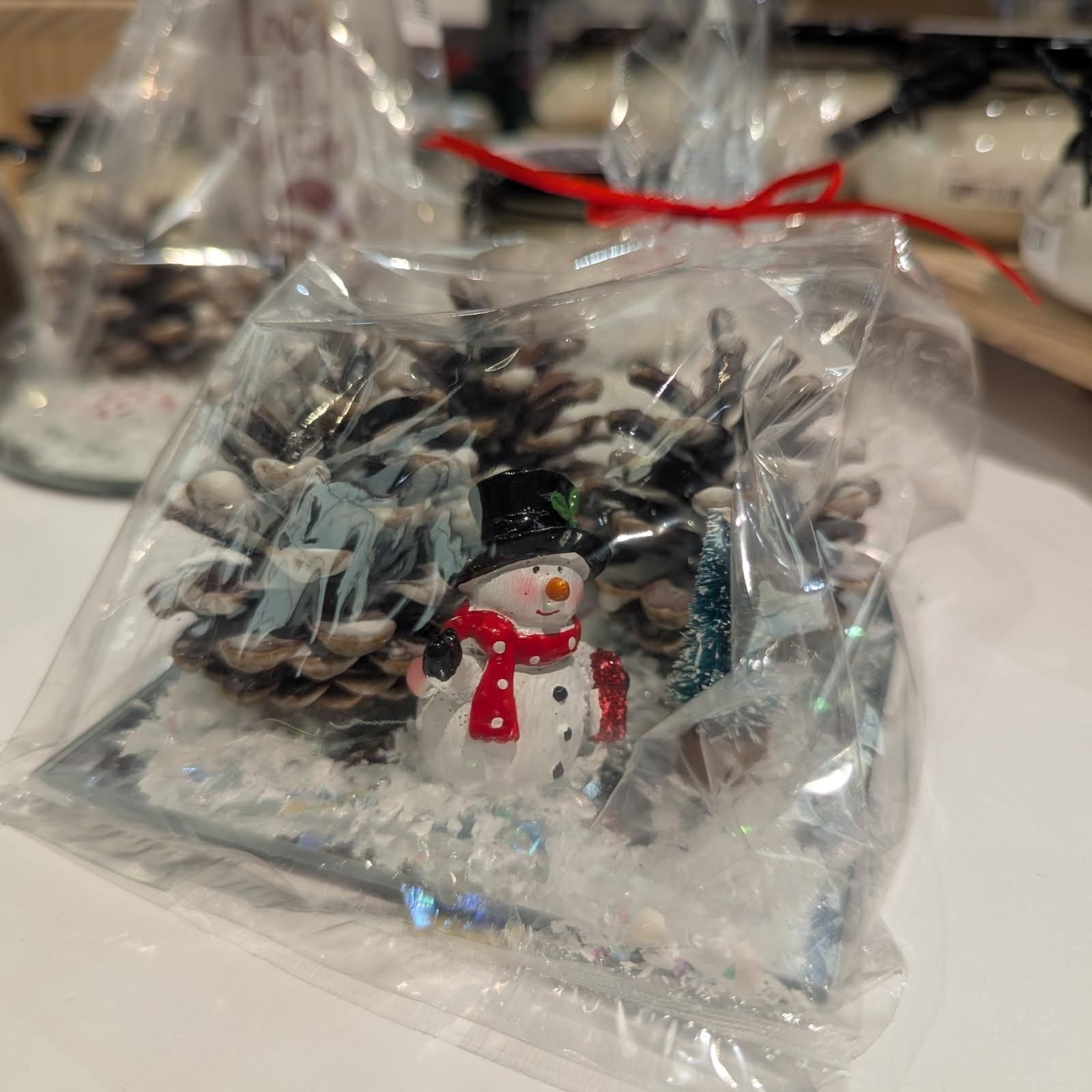 Snowman figurine in a winter scene, encased in clear plastic bag tied with a red ribbon, on a white surface.