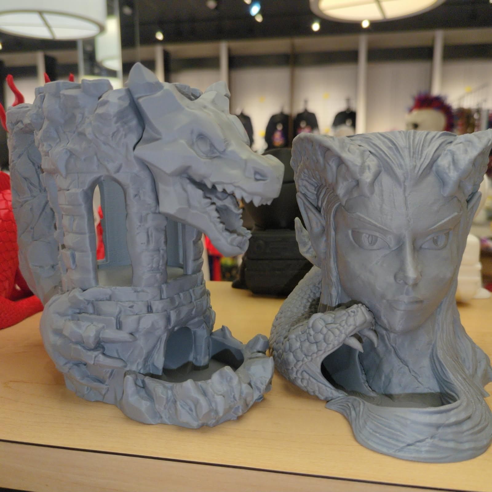 Two gray 3D-printed dice towers: a dragon head and a woman's face, displayed on a shelf.