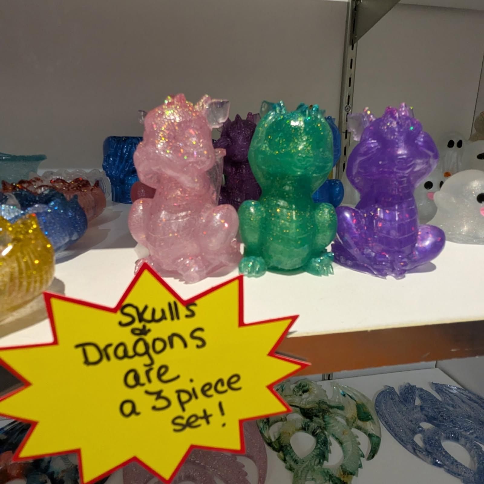 Three glittery dragon figurines, pink, green, and purple, on a shelf with a sign: 