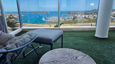 Lounge chairs on a balcony overlooking a blue ocean and coastal town through large windows