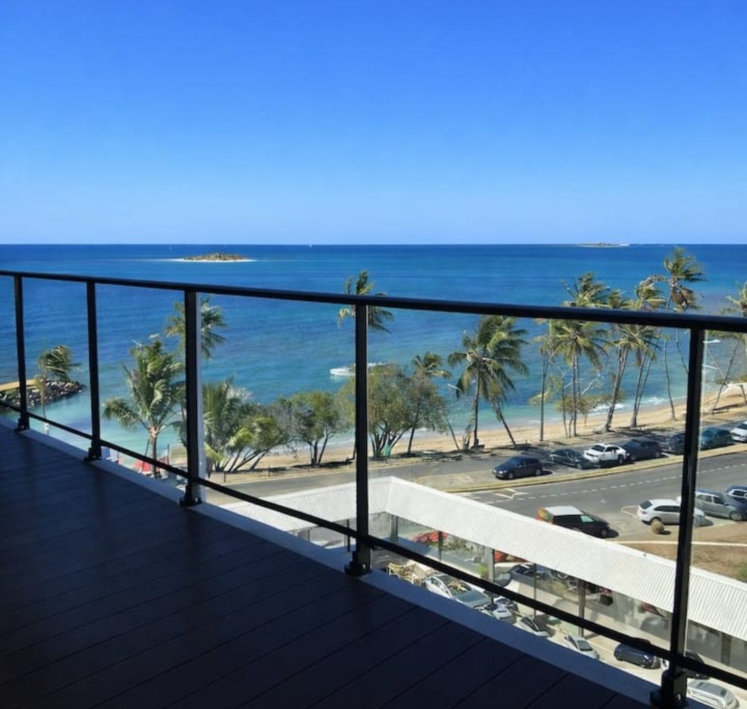 Balcony view of turquoise ocean, palm trees, beach, and parked cars under a clear blue sky