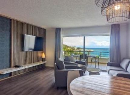 Modern hotel living room with TV, seating, and large window overlooking ocean views
