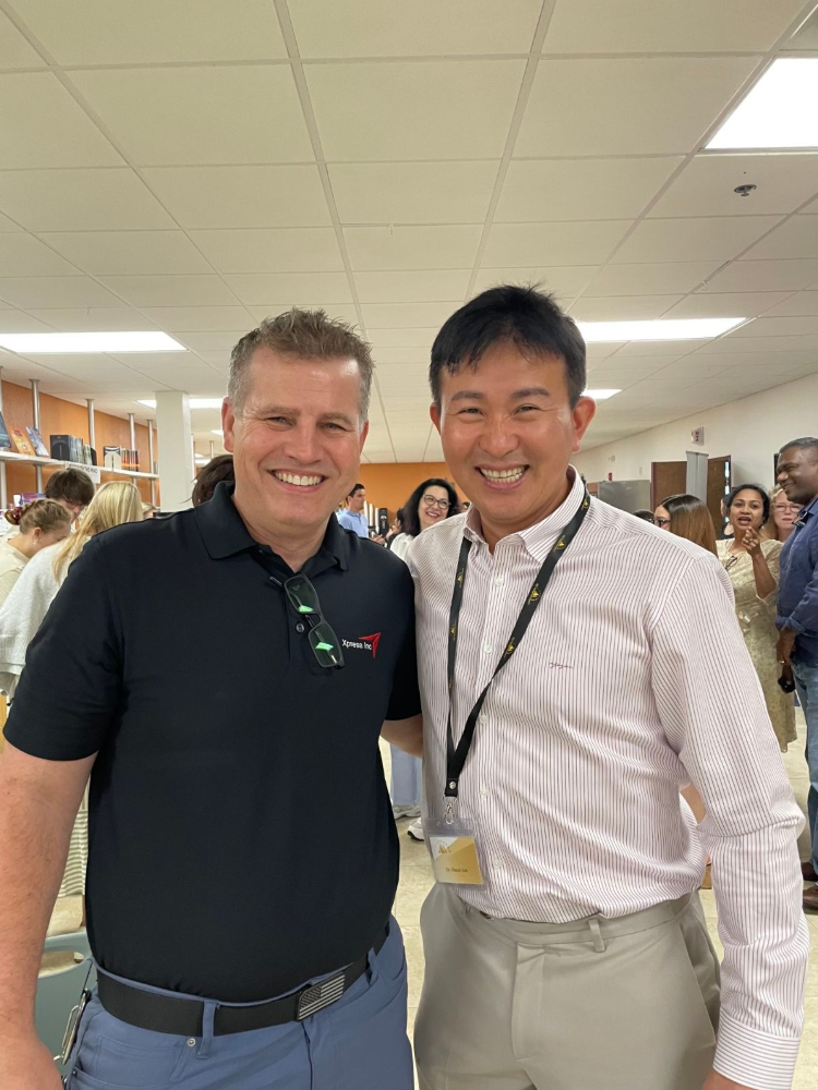 Two men pose smiling for a photo in a brightly lit hallway.