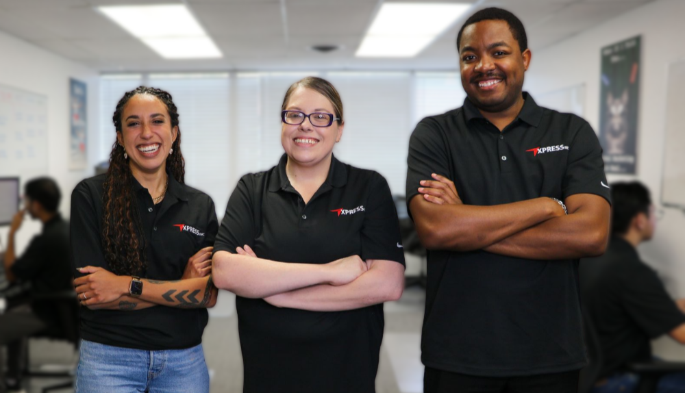 Three professionals in black company shirts stand smiling with arms crossed in a brightly lit office setting.