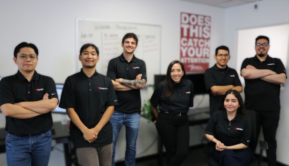 A group of seven colleagues in matching black polo shirts stand in an office, smiling for a team photo.