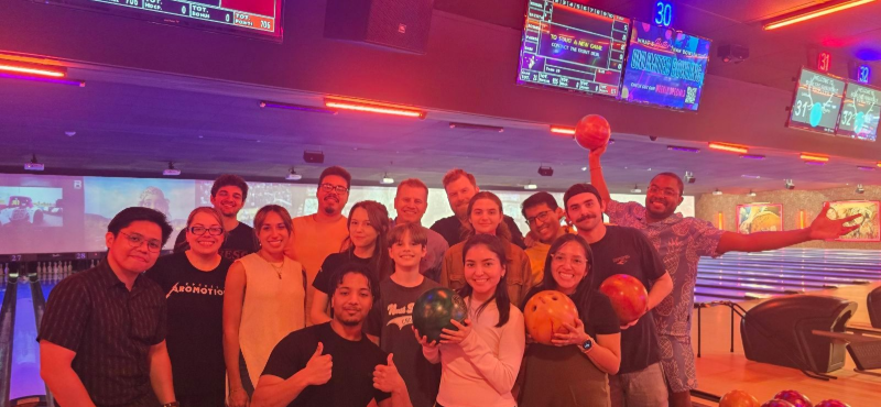 Group of people posing at a bowling alley, some holding bowling balls. Lit with red and purple lights.