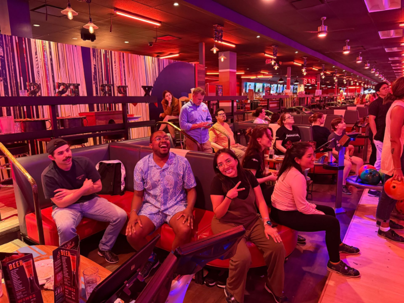 People at a bowling alley. Some seated, smiling, others standing nearby. Red and purple lighting.