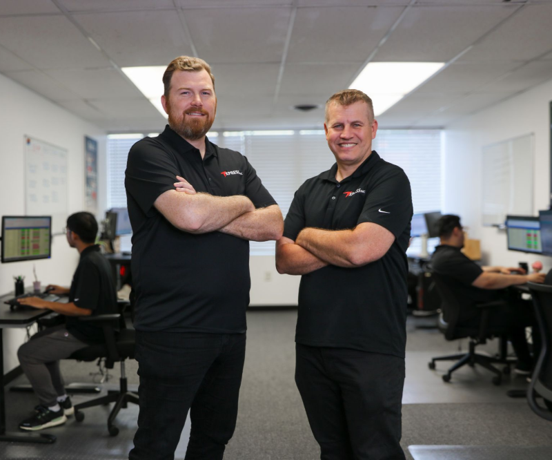 Two men in black shirts with arms crossed, standing in an office with other people working at computers.