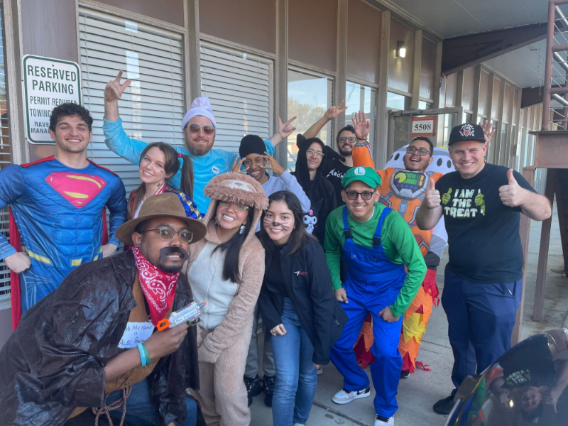 Group of people in costumes posing outside a building, some giving thumbs up or raising their arms.