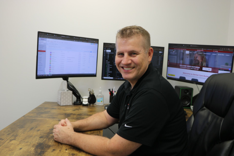 Man sitting at desk with three computer monitors, smiling.