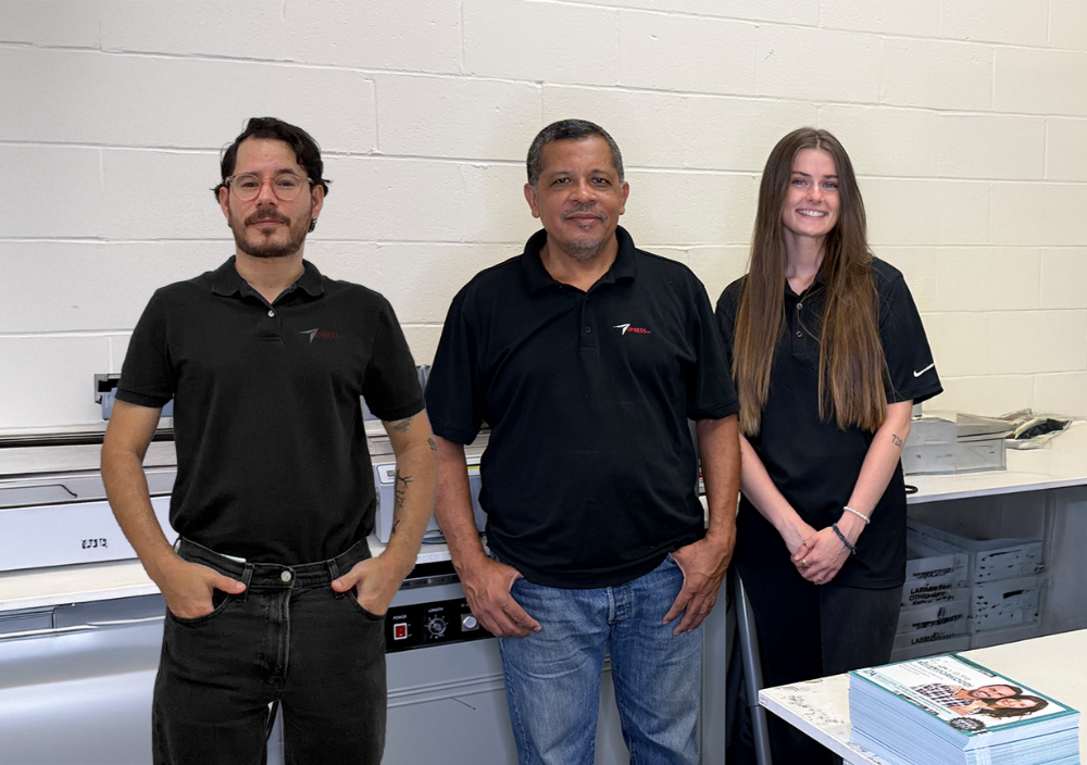 Three people in black shirts standing, posing near a printing machine.