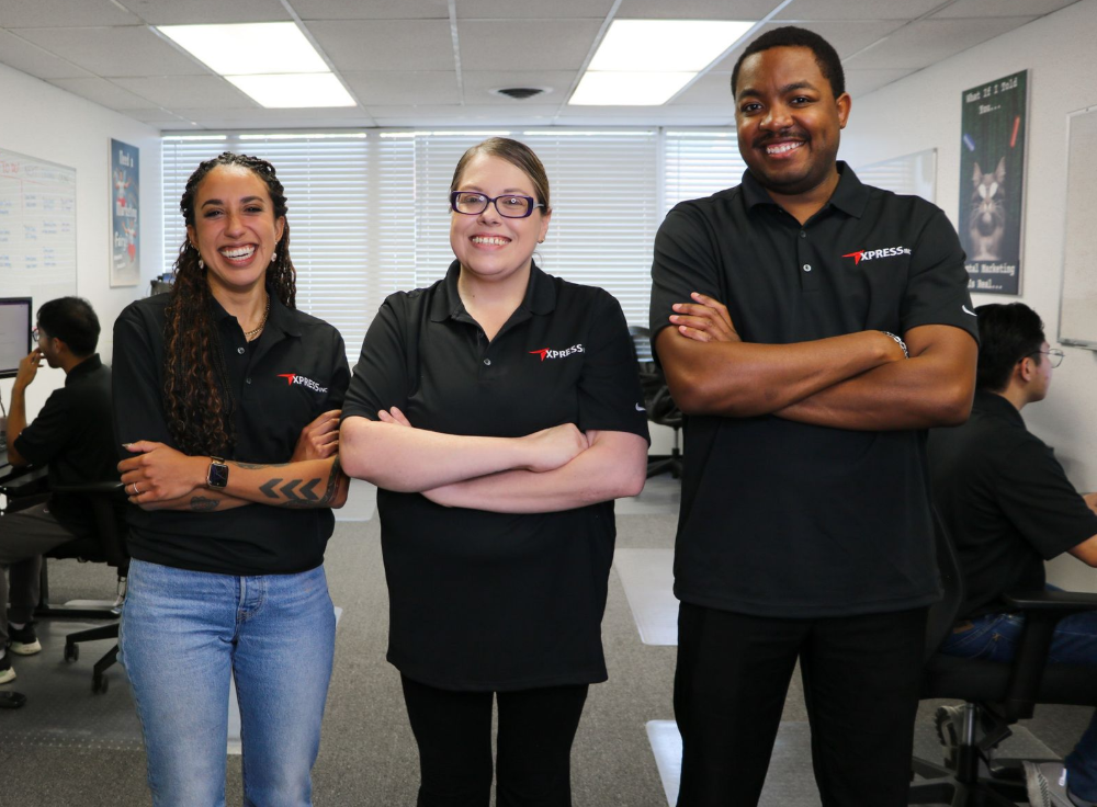 Three people in black shirts with arms crossed, standing in an office with other people in the background.