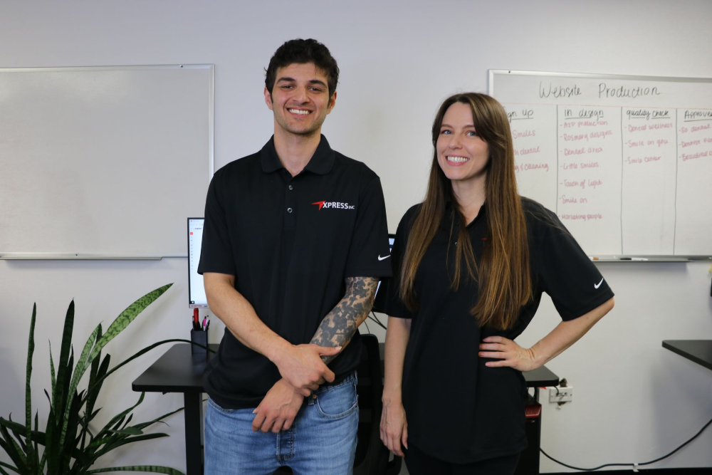 Two people in black shirts stand in front of a whiteboard and desk, smiling.