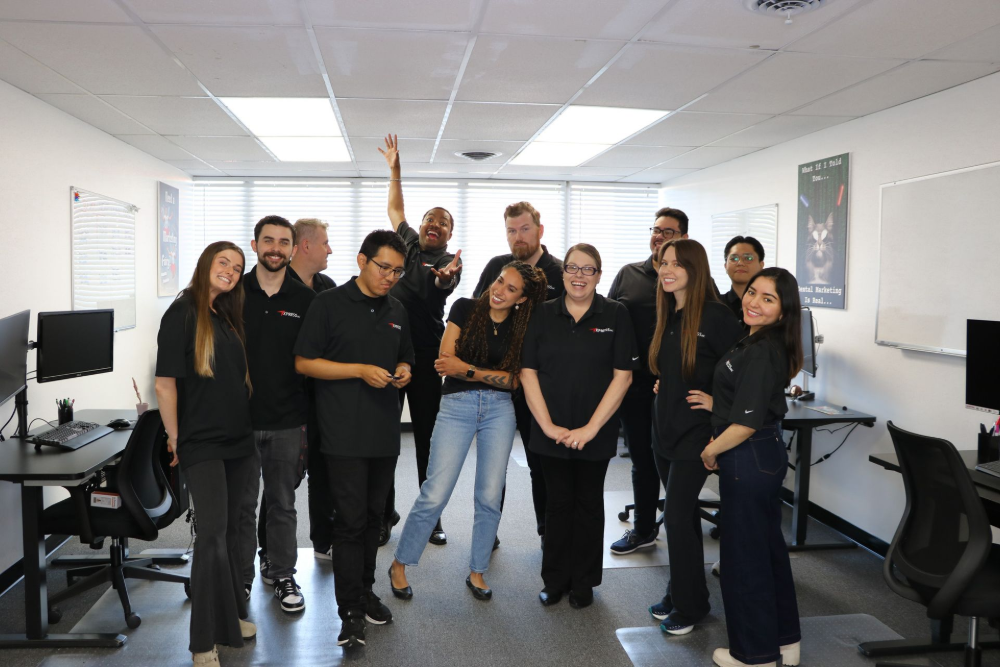 Group of people in black shirts pose in an office, smiling. Some are raising their arms in celebration.