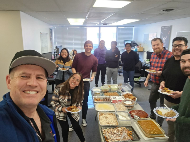 Office colleagues gather around a table laden with food, smiling. Some hold plates. Indoors with bright window.