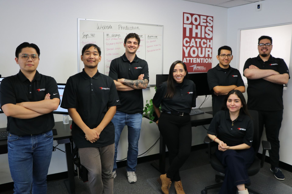 Group of people in black shirts posing in an office, white board in background. 