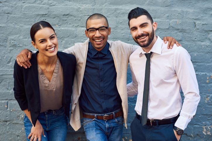 Three smiling people with arms around each other pose against a gray brick wall.