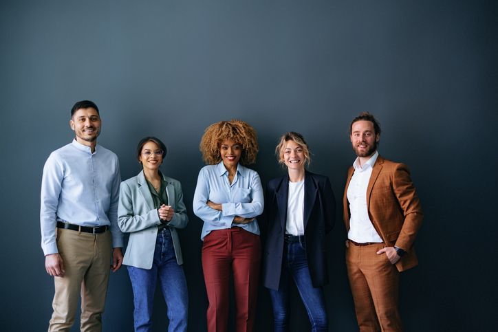 Five people in business attire smiling in front of a blue-gray wall.