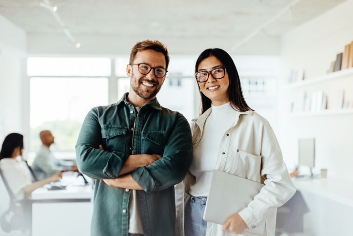 Two people smiling in office setting; man with arms crossed, woman holding laptop.