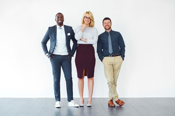 Three people standing against a white wall: a man in a suit, a woman in a skirt, and a man in a button-up shirt and khakis.